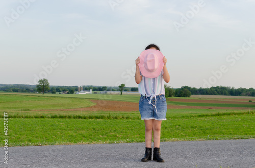 Girl covering her face with a cowboy hat in the field during sunset, wears a pink hat, surrounded by green grass.