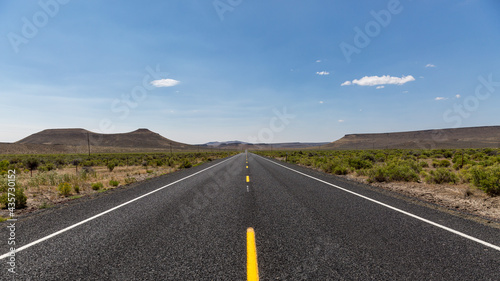 Straight asphalt road with dashed yellow strip line running between hills into horizon. A road in a high desert of South Oregon