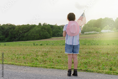 Young cowgirl with her back to the camera holding an American flag on the green grass of a farm during sunset, Independence Day concept.