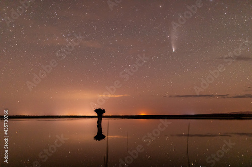 Panorama of a night sky with comet Neowise over a small lake in SW Oregon. Stars, clouds, and distant light reflect on a surface. Long exposure photo