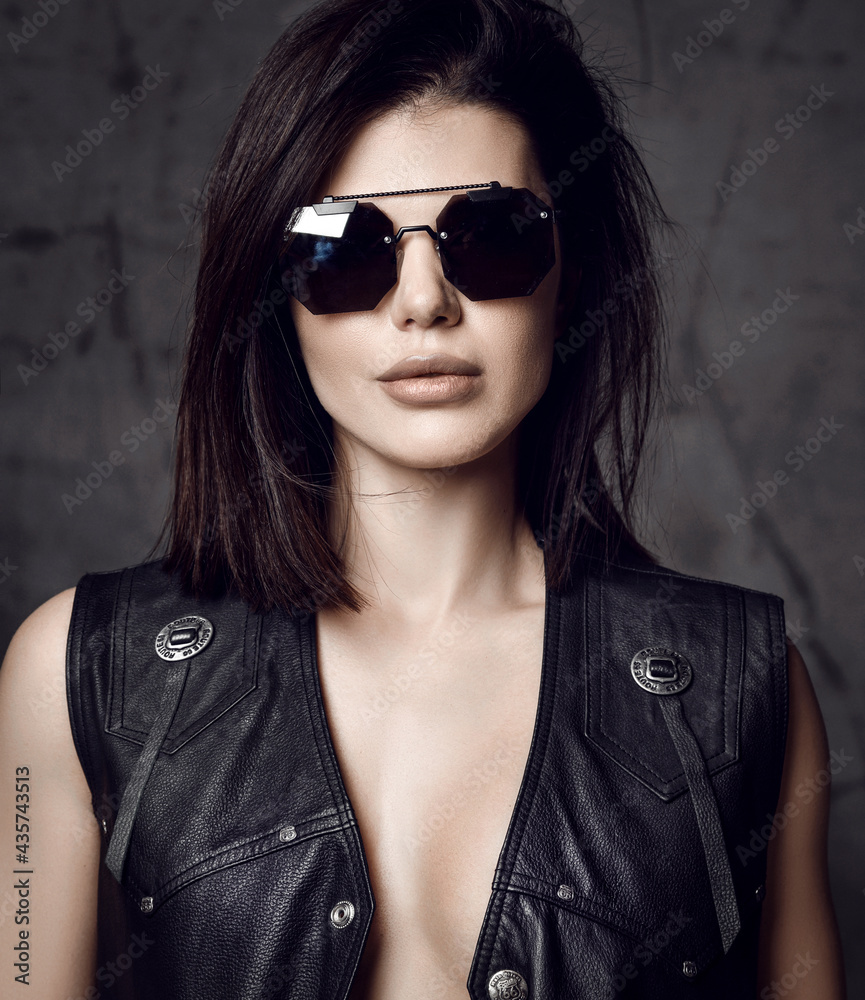 Portrait of powerful brutal sexy pretty brunette woman in black leather vest on naked body and sunglasses over grey concrete wall background pic