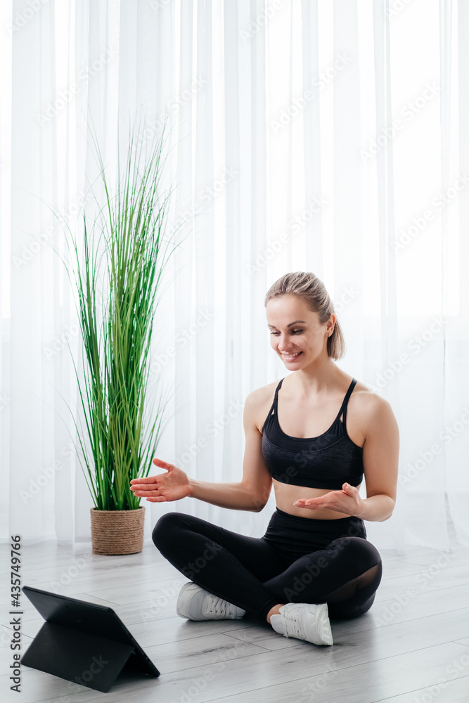 Online sport training. Female trainer. Fitness blog. Healthy body. Happy athletic woman in black sportswear in lotus pose on floor speaking to tablet computer light room interior. Online sport training. Female trainer. Fitness blog. Healthy body. Happy athletic woman in black sportswear in lotus pose on floor speaking to tablet computer light room interior.