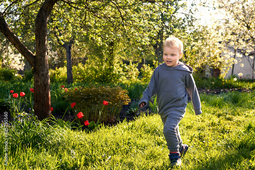 A three-year-old joyful boy runs on the grass in a blooming garden.