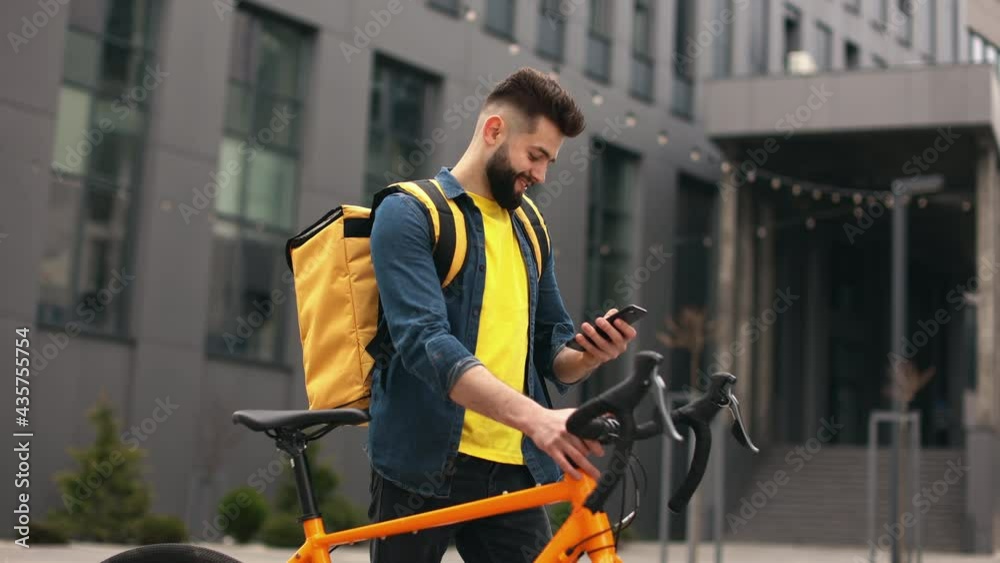 The food delivery guy is standing and smiling. He is looking at the smartphone. He is holding a bicycle and carrying a large backpack on his shoulders. The camera is moving around. Beautiful Business The food delivery guy is standing and smiling. He is looking at the smartphone. He is holding a bicycle and carrying a large backpack on his shoulders. The camera is moving around. Beautiful Business