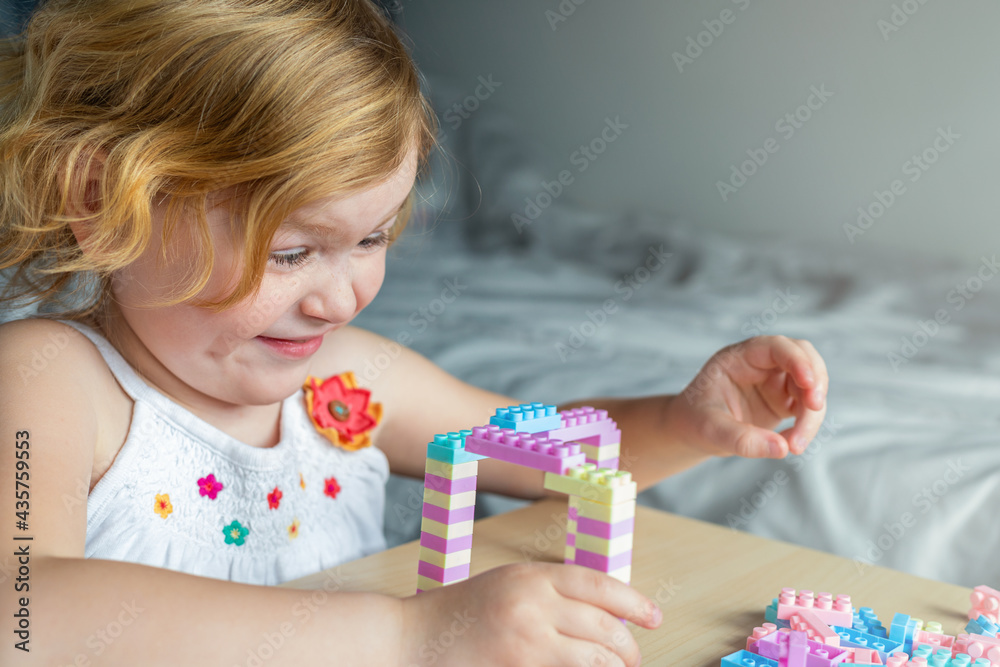 Smiling happy little beautiful girl playing with toy plastic building blocks, sitting at the table. Small child busy with fun creative leisure activity. Development of fine motor skills for children. Smiling happy little beautiful girl playing with toy plastic building blocks, sitting at the table. Small child busy with fun creative leisure activity. Development of fine motor skills for children.