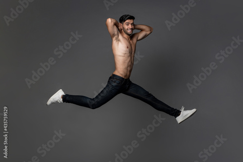 Fototapeta Naklejka Na Ścianę i Meble -  Handsome shirtless smiling Caucasian man jumping with legs open wide in gray isolated studio background