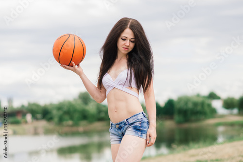 Young athletic female, in top and sweatpants, playing with ball on basketball court outdoors.