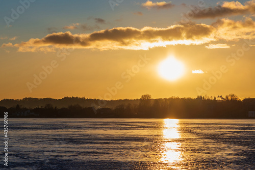 Winter landscape with frozen lake at sunrise or sunset. Lake glistening ice reflect a sun.Forest in the background.