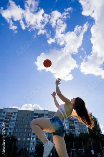 Young athletic female, in top and sweatpants, playing with ball on basketball court outdoors.