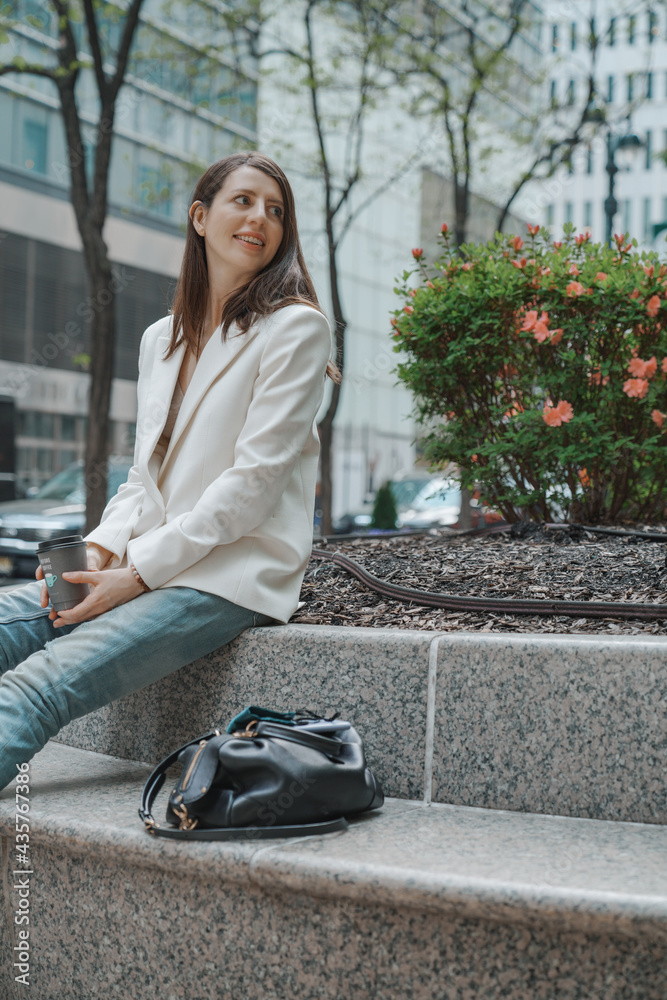 Smiling positive young woman relaxing having a break, city streets. Stylish fashion blogger concept, long brown hair, blazer, blue jeans, leather bag. New York modern buildings background Smiling positive young woman relaxing having a break, city streets. Stylish fashion blogger concept, long brown hair, blazer, blue jeans, leather bag. New York modern buildings background