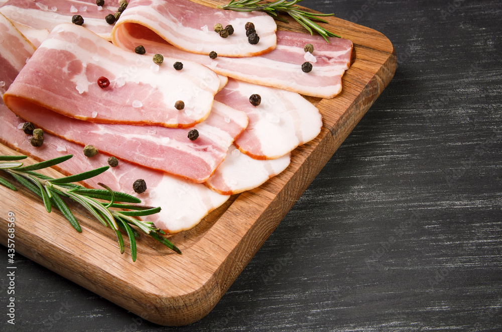 Board with raw bacon with spices, and rosemary on a black background Board with raw bacon with spices, and rosemary on a black background