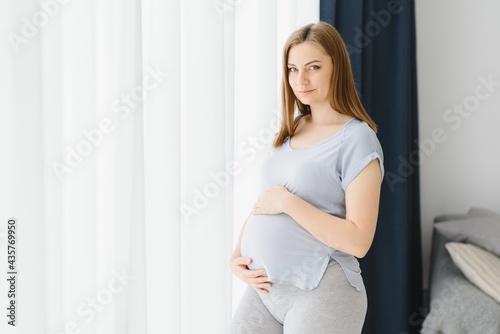 Beautiful pregnant woman is touching her belly and smiling while standing near the window at home