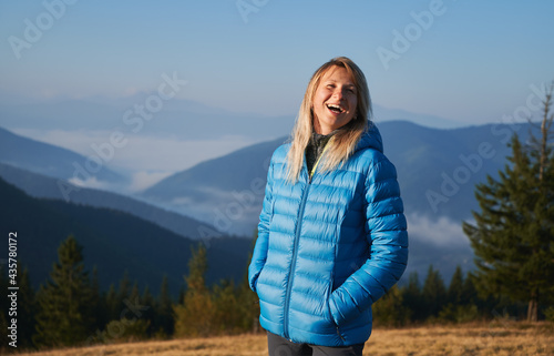 Portrait of a happy woman standing on background of mountain hills covered with fog and smiling. Walk in the fresh mountain air under blue sunshine sky in the morning.
