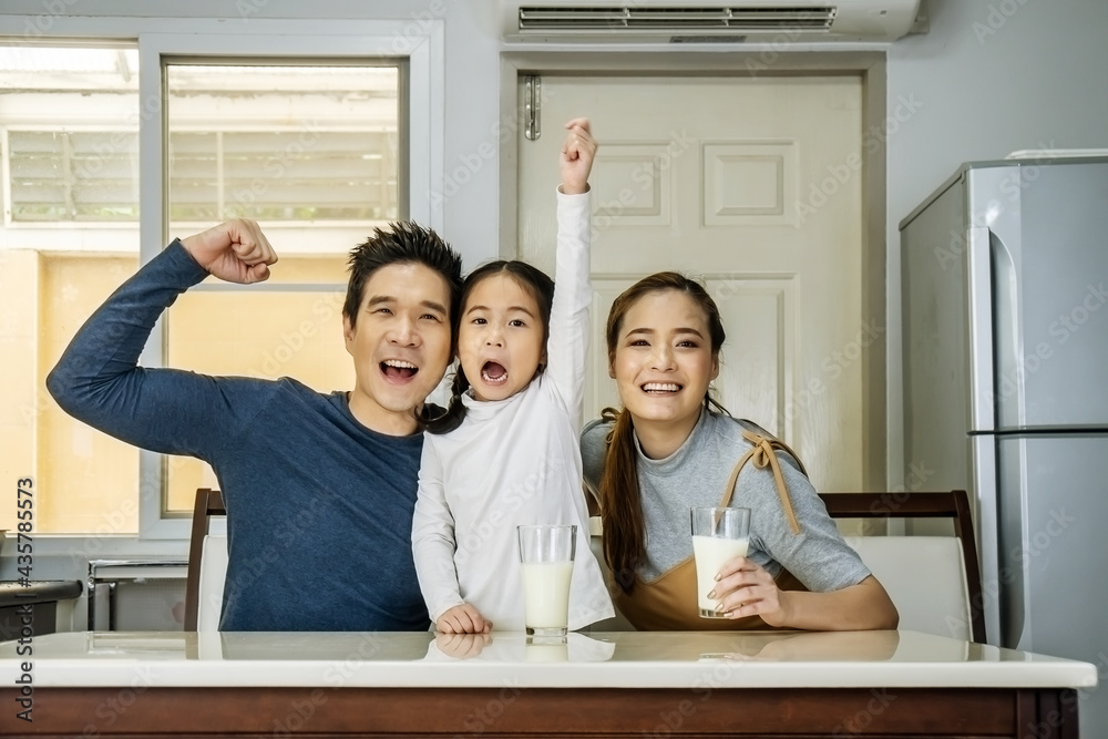Fototapeta premium Happy family having fun in the kitchen. Asian Father, mother and little daughter spending time together and having breakfast drinking and hold glasses of milk at table
