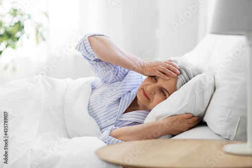 health, old age and people concept - senior woman in pajamas suffering from headache lying in bed at home bedroom