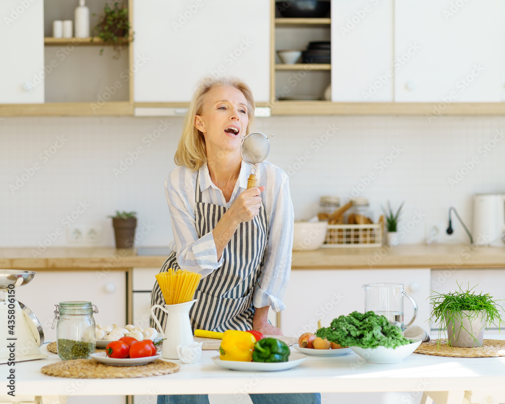 Playful carefree senior woman in apron using metal strainer as microphone and singing, having fun while cooking healthy food in modern light kitchen at home. Happy mature female enjoying retired life