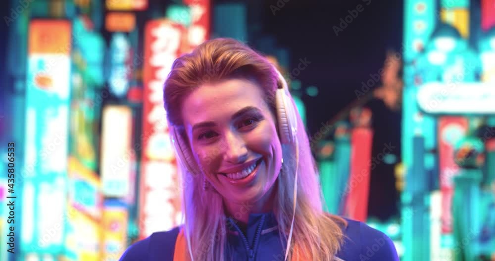 Portrait view of the blonde caucasian woman wearing headphones posing at the camera while listening music at the street with neon buildings at the background Portrait view of the blonde caucasian woman wearing headphones posing at the camera while listening music at the street with neon buildings at the background