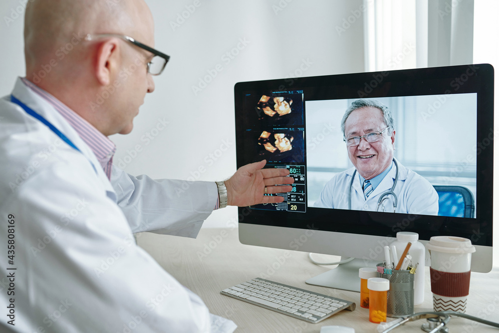 Over shoulder view of mature Caucasian doctor in lab coat pointing at data on computer while discussing medical research with colleague via videoconferencing