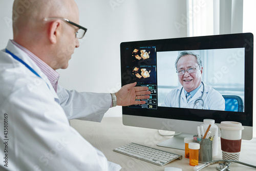 Over shoulder view of mature Caucasian doctor in lab coat pointing at data on computer while discussing medical research with colleague via videoconferencing © DragonImages