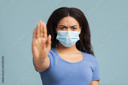 Social distance concept. Serious african american lady in protective mask showing stop gesture against blue background