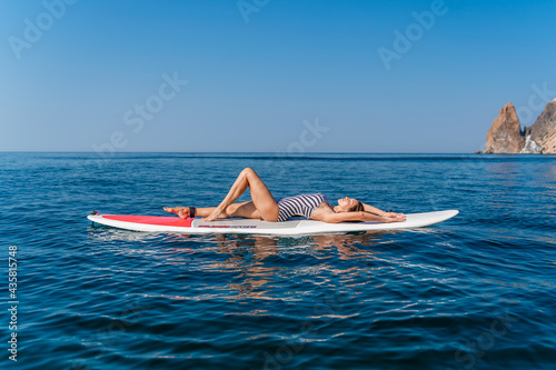 Sporty girl on a glanders surfboard in the sea on a sunny summer day. In a striped swimsuit, he lies on his back. Summer activities by Stortom by the sea