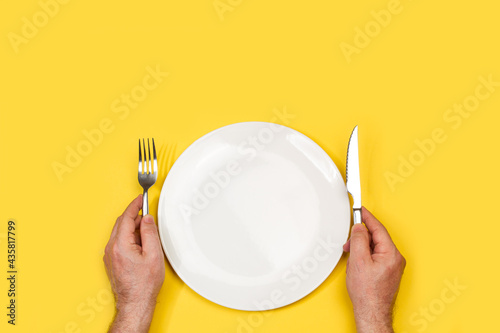 Manos de hombre sosteniendo unos cubiertos junto a un plato redondo blanco vacío sobre un fondo amarillo liso y aislado. Vista superior. Copy space photo