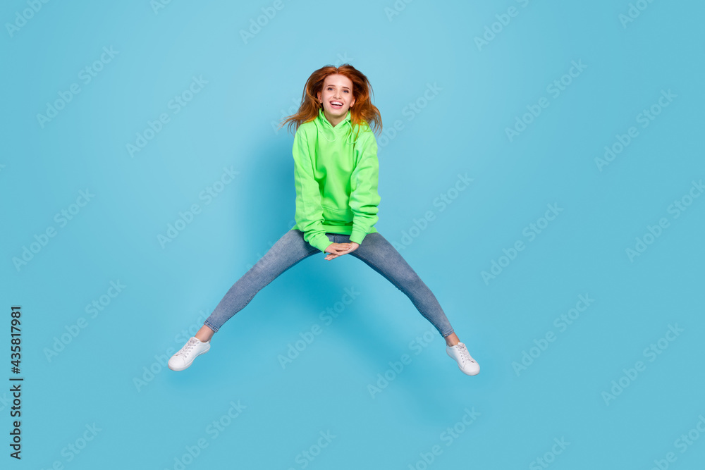 Photo of carefree funny young woman wear green sweatshirt jumping high smiling isolated blue color background