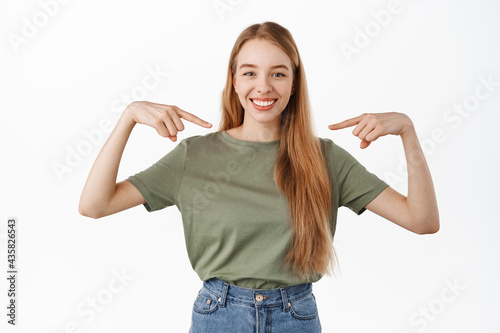 Confident blond girl smiles with white perfect teeth, pointing at herself and looking determined at camera, self-promoting, volunteering, pick me choose her gesture, standing over white background photo