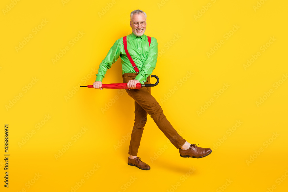 Full length profile side photo of senior man happy smile have fun go walk umbrella weather isolated over yellow color background Full length profile side photo of senior man happy smile have fun go walk umbrella weather isolated over yellow color background
