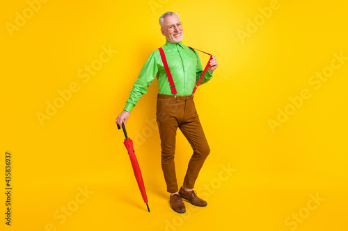 Full size photo of senior man happy positive smile hand touch suspenders parasol forecast isolated over yellow color background