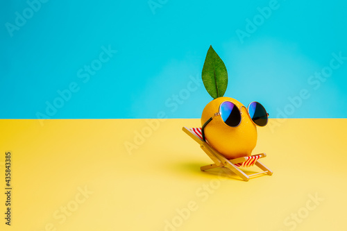 Lemon fruit chilling in beach chair on the blue and yellow background. Summer vacation concept. Sunglasses on lemon with green leaf relaxing on the sunbed. Creative art, minimal aesthetic.