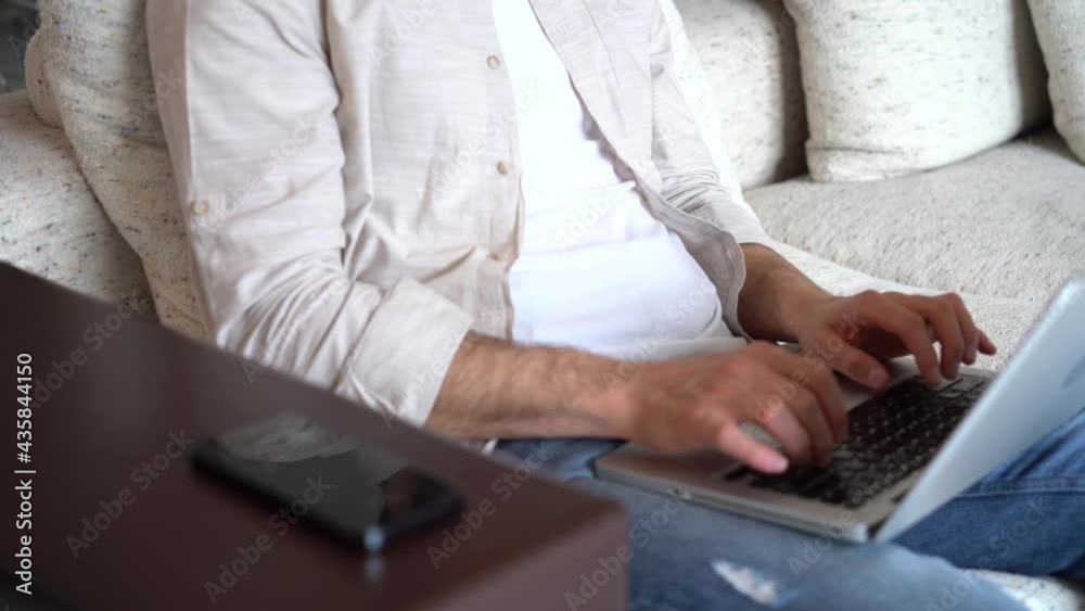 Young man talking on smartphone while sitting on couch with opened laptop. Responsible company member working remotely at home office. Young man talking on smartphone while sitting on couch with opened laptop. Responsible company member working remotely at home office.