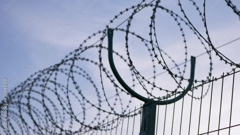 Barbed wire Hanging on the Border of Iron Fence on Against the Blue Sky. Restricted area. Barrier from strangers people, refugees, prisoners. Lattice. Jail. 4K. Close up. Barbed wire Hanging on the Border of Iron Fence on Against the Blue Sky. Restricted area. Barrier from strangers people, refugees, prisoners. Lattice. Jail. 4K. Close up.