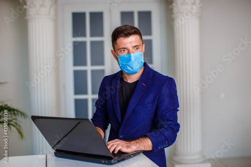 A young man in a medical mask and a blue jacket works on a laptop in a brightly lit room. internet office