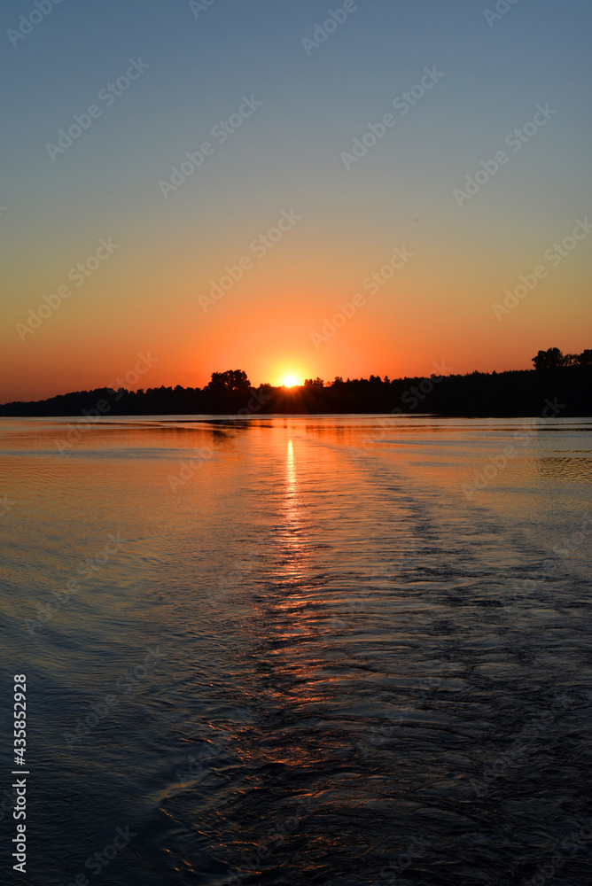Naklejka premium Early morning. Sunrise. View from the river with the reflection of the shore in the water and the wake of the ship. Vertical