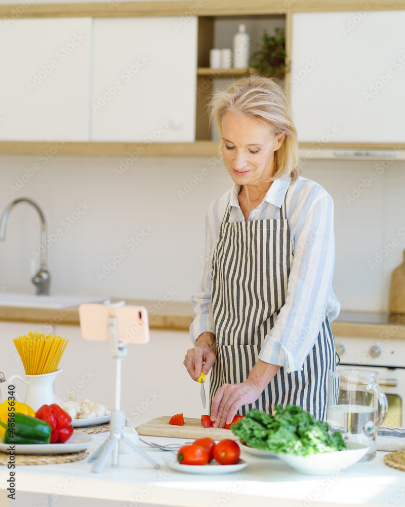 Vertical shot of cheerful senior food blogger in apron recording video for culinary blog on smartphone, showing process of cooking healthy food in modern light kitchen, cutting fresh vegetables