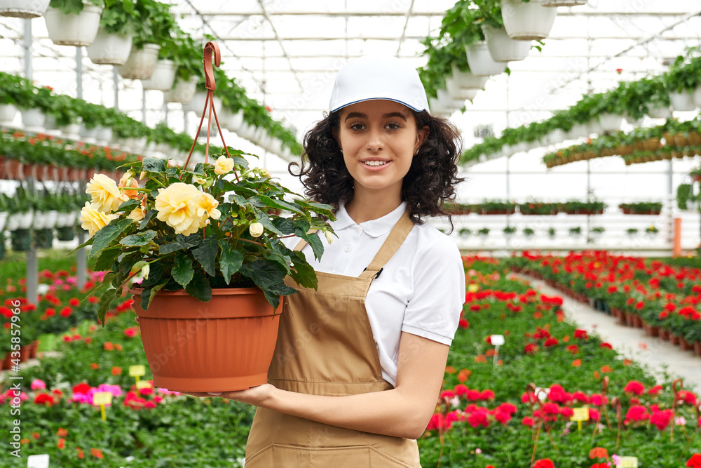 Portrait of attractive young woman in white cap and beige apron posing at greenhouse with flower pot in hands. Concept of people, work and horticulture.