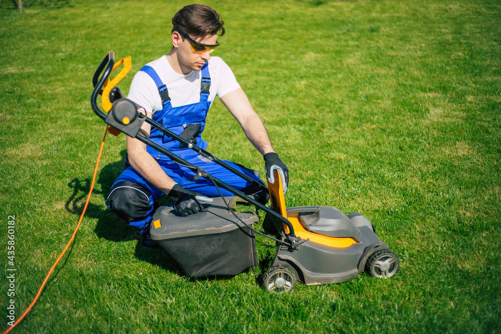Young man mows the lawn using an electric lawn mower in a special worker suit near a large country house in the backyard Young man mows the lawn using an electric lawn mower in a special worker suit near a large country house in the backyard