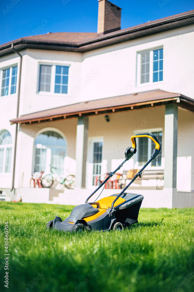 Photo of a modern electric lawn mower standing on a backyard lawn near a house Photo of a modern electric lawn mower standing on a backyard lawn near a house
