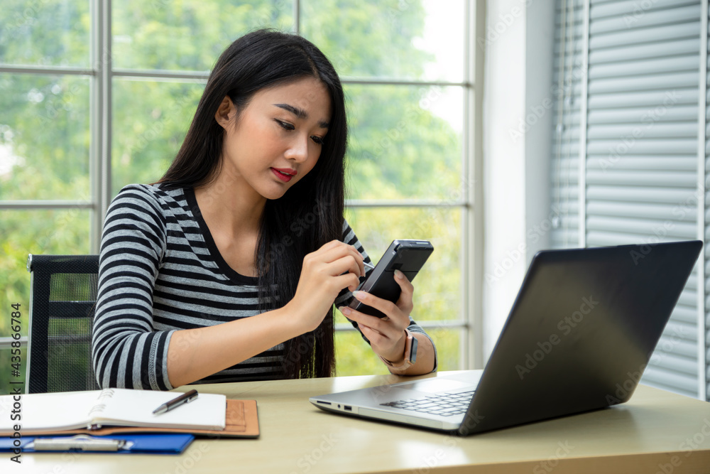 Happy young Asian woman smiling writing memo on pocket PC taking data from laptop in living room. teenage work from home concept Happy young Asian woman smiling writing memo on pocket PC taking data from laptop in living room. teenage work from home concept