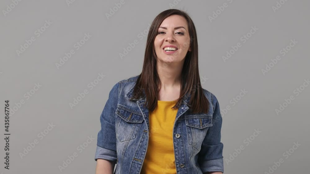 Smiling fun beautiful brunette young woman 20s years old in denim jacket yellow t-shirt posing looking camera charming smile isolated on grey color background studio. People emotions lifestyle concept Smiling fun beautiful brunette young woman 20s years old in denim jacket yellow t-shirt posing looking camera charming smile isolated on grey color background studio. People emotions lifestyle concept