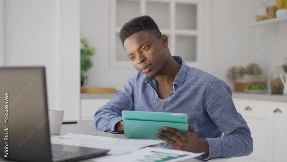 Portrait of young handsome man scrolling tablet screen checking laptop sitting at kitchen table indoors. African American confident freelancer surfing Internet analyzing e-market in home office Portrait of young handsome man scrolling tablet screen checking laptop sitting at kitchen table indoors. African American confident freelancer surfing Internet analyzing e-market in home office