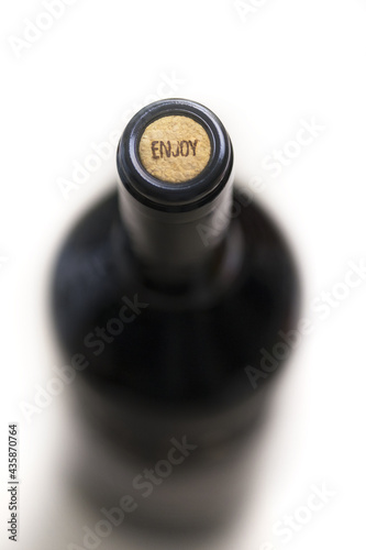 Top close view of unopened dark glass bottle of red wine with cork with enjoy sign on it on white background. Selective focus, blurred background. Wine culture, advertisement, marketing concept.