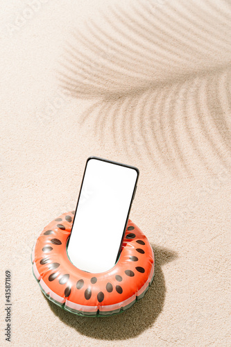 Inflatable watermelon shaped with a mobile phone inside on tropical beach sand with shadows of coconut tree leaves in summer. Travel and holiday concept background.vertical image with copy space