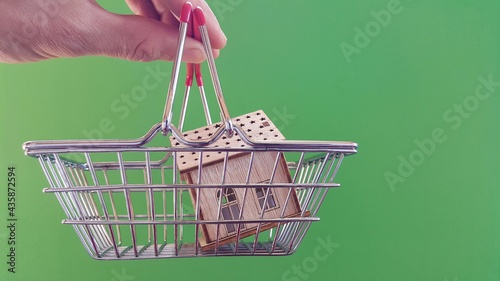 Real estate market and real estate purchase, the fingers of a person holding a house icon and a shopping cart. Buying a home concept on a green background. Copy space. photo