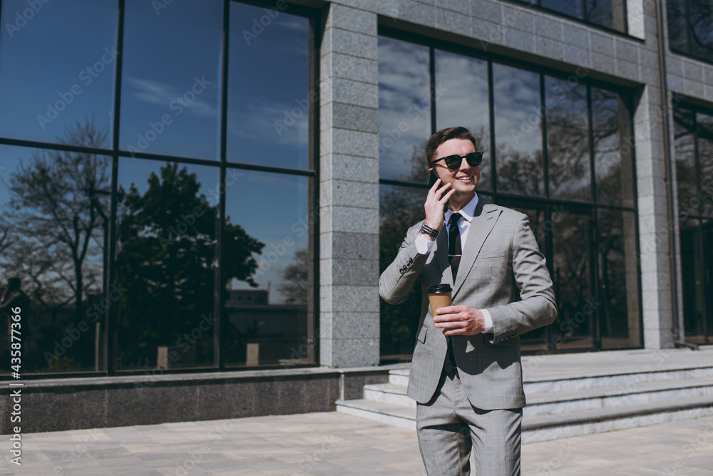 Young happy employee business man 20s in grey suit walk near office glass wall building outdoors in downtown city center hold mobile cell phone takeaway craft paper brown cup coffee to go look aside. Young happy employee business man 20s in grey suit walk near office glass wall building outdoors in downtown city center hold mobile cell phone takeaway craft paper brown cup coffee to go look aside.