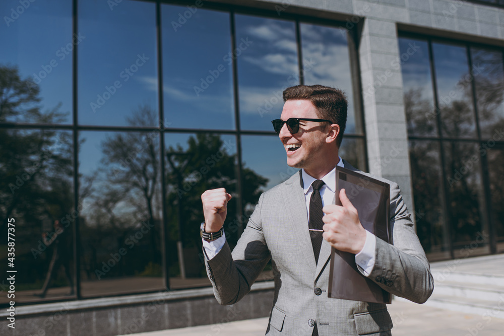 Young employee business man 20s in grey suit walk go near office glass wall building outdoors in downtown city center hold black folder for papers document do winner gesture clench fist look aside Young employee business man 20s in grey suit walk go near office glass wall building outdoors in downtown city center hold black folder for papers document do winner gesture clench fist look aside