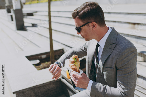Young european caucasian businessman man in grey formal suit with tie eyeglasses sitting on concrete steps in city center eating biting croissant have break lunch breakfast outdoors  look at watch
