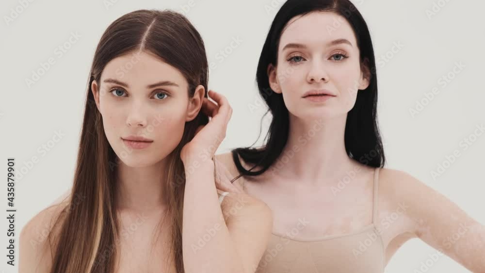 The two confident girls in knitted tops posing and straightening hair while standing in a white studio The two confident girls in knitted tops posing and straightening hair while standing in a white studio