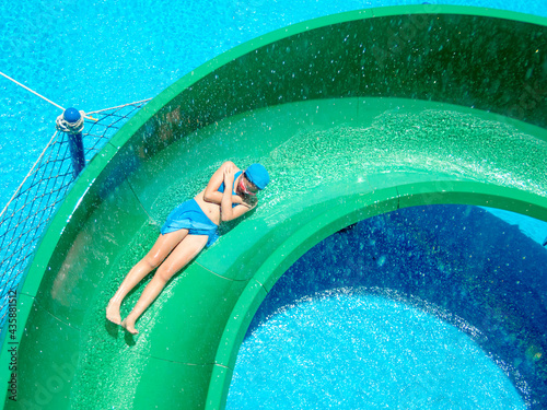 A hot summer day, on a swimming pool, an Asian young girl in a swimming suit is slipping down a green waterslide